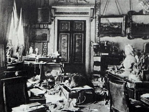 A black and white photograph of a study with papers, books and broken furniture strewn across the floor