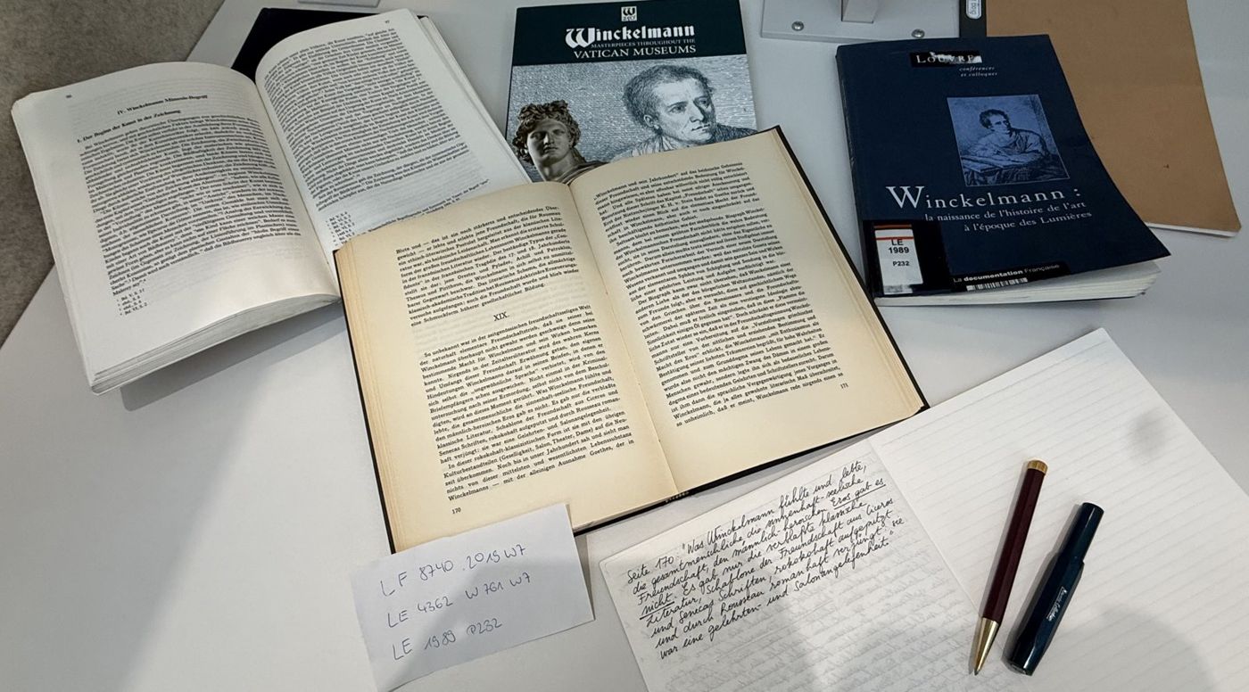 Open books on Winckelmann on a library table, with a handwritten page of notes in German and two pens